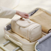 Mano che posiziona un beauty case beige in una valigia aperta. Organizzatore da viaggio, borsa da toilette, accessori da viaggio essenziali.