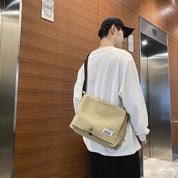 Uomo con borsa a tracolla beige in tessuto, indossa cappello nero e maglia bianca, in un ambiente moderno con ascensori.