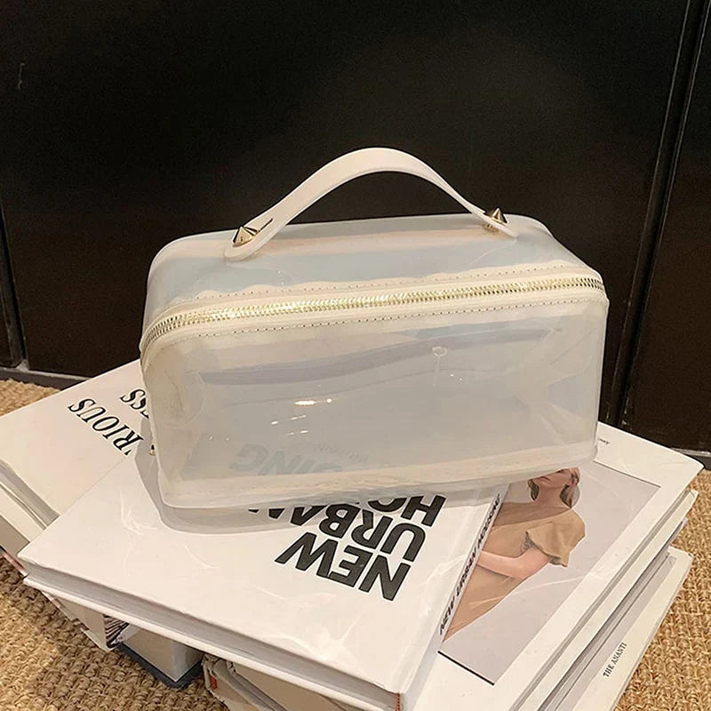 Borsa trasparente in PVC con manico bianco e cerniera dorata, posizionata su libri. Elegante e pratica per viaggi e organizzazione.