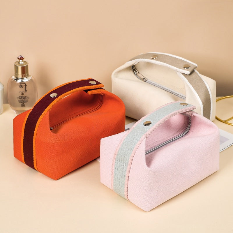 Tre borse cosmetiche in tessuto colorato (arancione, rosa, beige) con cerniera e manico, su sfondo beige. Accessori da viaggio eleganti e pratici.