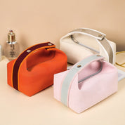 Tre borse cosmetiche in tessuto colorato (arancione, rosa, beige) con cerniera e manico, su sfondo beige. Accessori da viaggio eleganti e pratici.