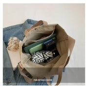 Borsa tote beige in tela con manici, contenente un ombrello, un power bank e un tablet. Sfondo con giacca di jeans. Accessori moda pratici.