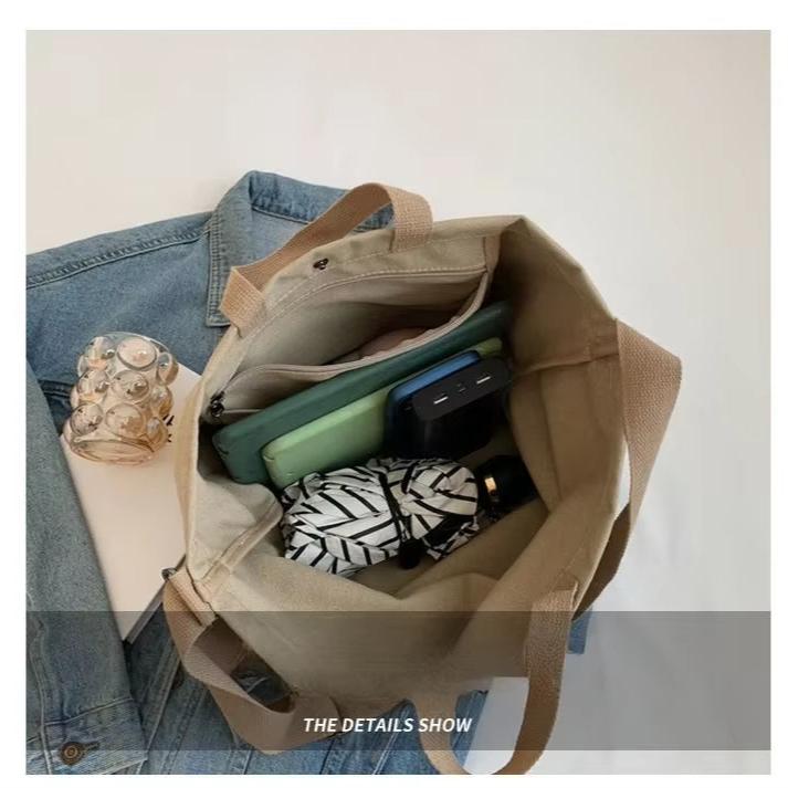 Borsa tote beige in tela con manici, contenente un ombrello, un power bank e un tablet. Sfondo con giacca di jeans. Accessori moda pratici.