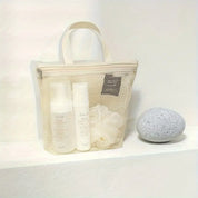 Borsa trasparente in rete con prodotti per la cura della pelle, flaconi bianchi e spugna, su sfondo bianco. Accessori da bagno eleganti e pratici.