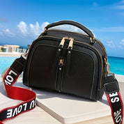 Borsa a tracolla nera in pelle con dettagli dorati, manico superiore e cinturino rosso con scritta "LOVE", posizionata su tavolo vicino a piscina.