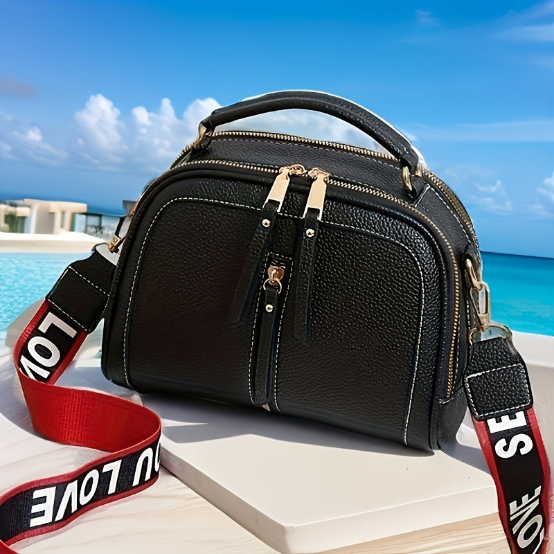 Borsa a tracolla nera in pelle con dettagli dorati, manico superiore e cinturino rosso con scritta "LOVE", posizionata su tavolo vicino a piscina.