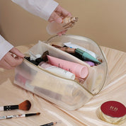 Borsa trasparente per cosmetici con cerniera, contenente creme, trucchi e pennelli su sfondo beige. Ideale per organizzare prodotti di bellezza.