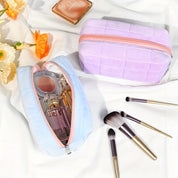 Due beauty case in tessuto pastello con zip, uno azzurro e uno viola, contengono profumi e cosmetici. Pennelli trucco e fiori decorativi accanto.