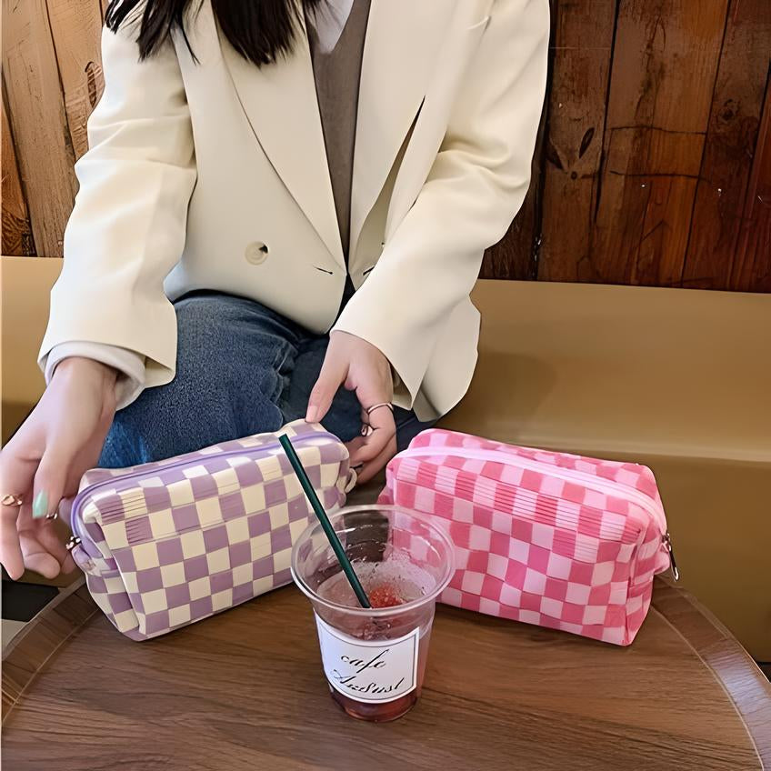 Donna con giacca bianca e jeans, tiene pochette a scacchi viola e rosa su tavolo di legno, accanto a bevanda rossa in bicchiere di plastica.