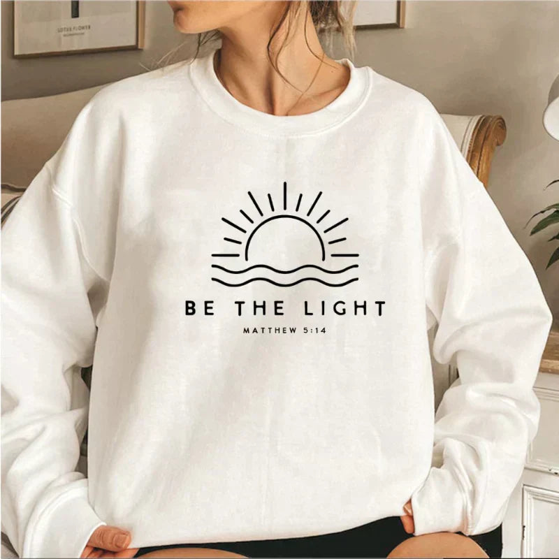 Donna con felpa bianca, stampa sole e scritta "Be the Light Matthew 5:14". Moda casual, abbigliamento ispirato alla fede, stile confortevole.