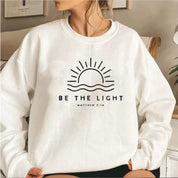Donna con felpa bianca, stampa sole e scritta "Be the Light Matthew 5:14". Moda casual, abbigliamento ispirato alla fede, stile confortevole.