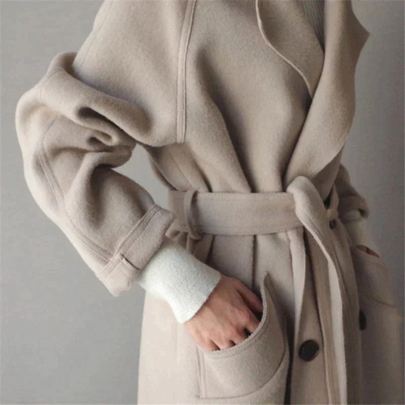 Cappotto beige in lana con cintura, tasche frontali e bottoni, indossato su maglione bianco. Elegante abbigliamento invernale donna.