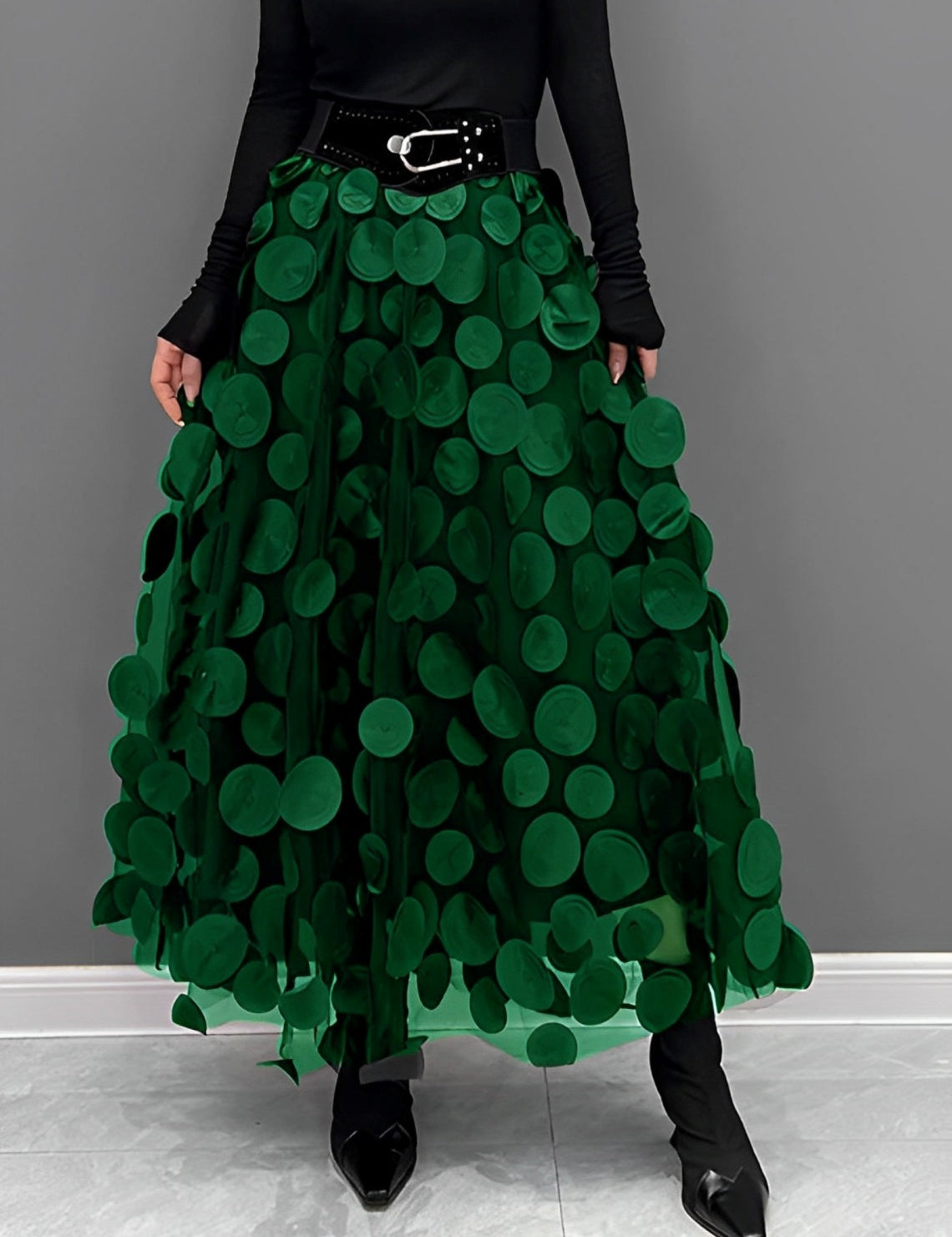 Gonna lunga verde con applicazioni circolari, cintura nera con fibbia, indossata con top nero a maniche lunghe e stivali a punta.