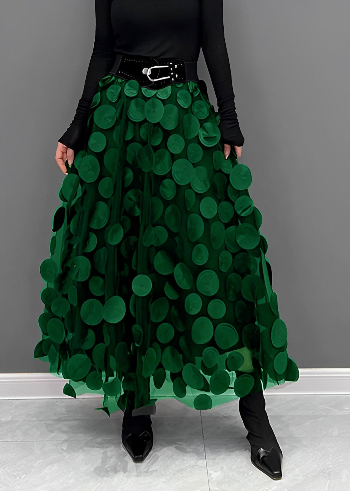 Gonna lunga verde con applicazioni circolari, cintura nera con fibbia, indossata con top nero a maniche lunghe e stivali a punta.