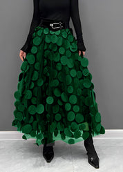 Gonna lunga verde con applicazioni circolari, cintura nera con fibbia, indossata con top nero a maniche lunghe e stivali a punta.