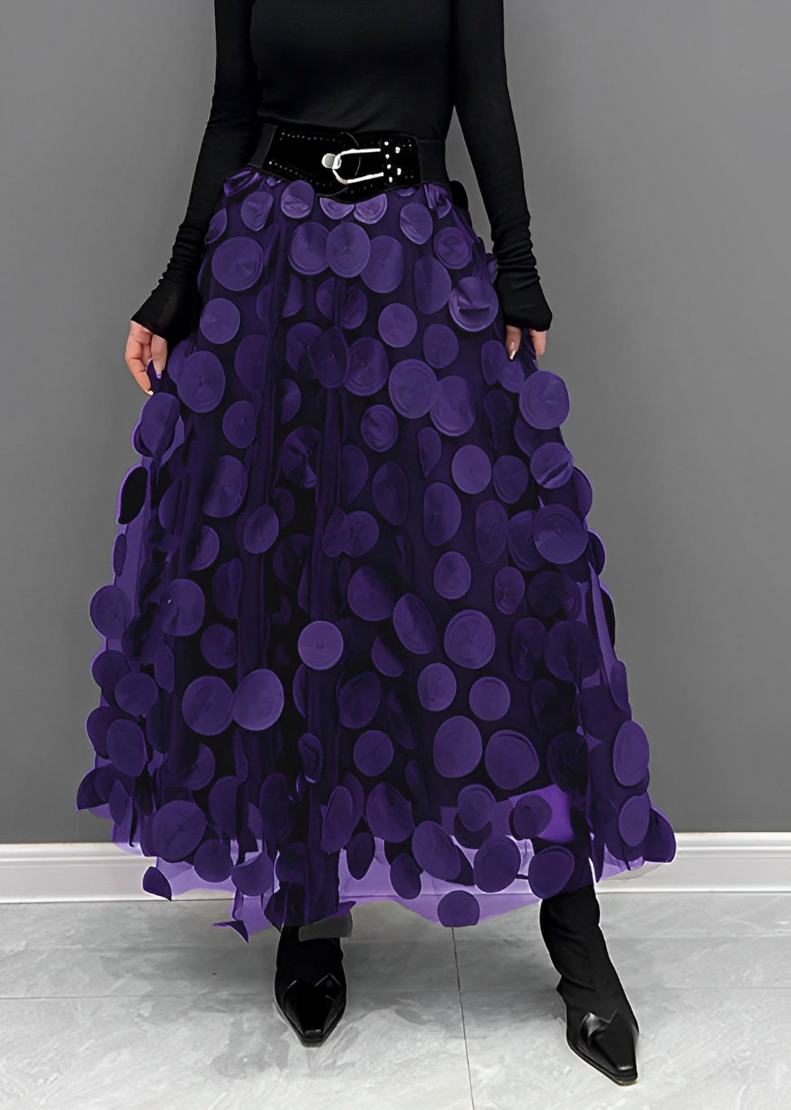 Gonna lunga viola con cerchi decorativi, cintura nera, indossata con maglia nera a maniche lunghe e stivali neri. Moda elegante donna.