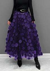 Gonna lunga viola con cerchi decorativi, cintura nera, indossata con maglia nera a maniche lunghe e stivali neri. Moda elegante donna.
