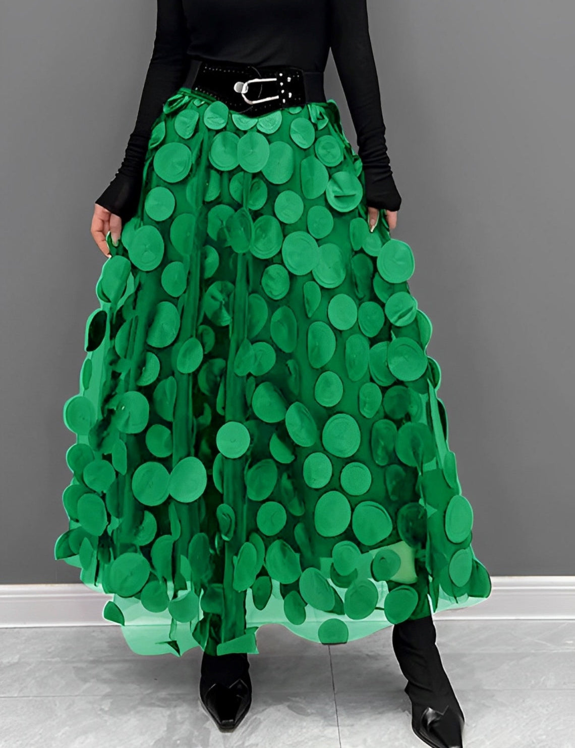Gonna lunga verde con cerchi decorativi, cintura nera, indossata con top nero a maniche lunghe e stivali a punta. Moda donna elegante e moderna.