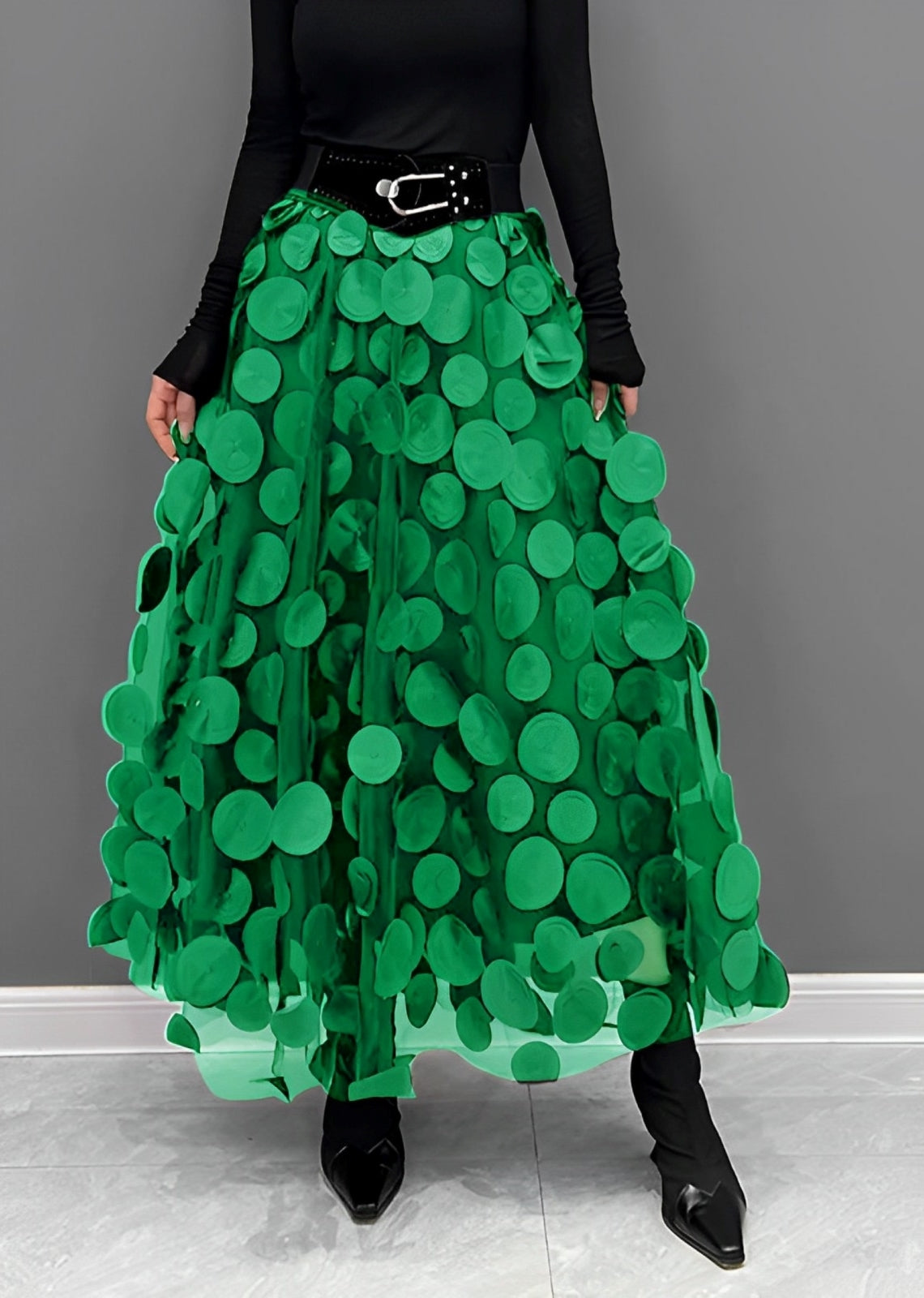 Gonna lunga verde con cerchi decorativi, cintura nera, indossata con top nero a maniche lunghe e stivali a punta. Moda donna elegante e moderna.
