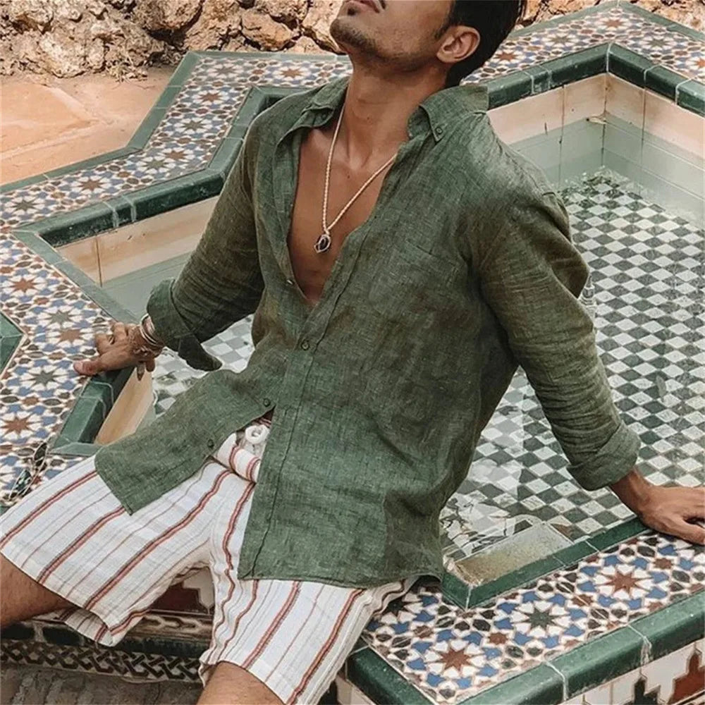 Uomo con camicia di lino verde e pantaloncini a righe bianche e marroni, seduto su fontana decorata con piastrelle geometriche. Moda estiva casual.