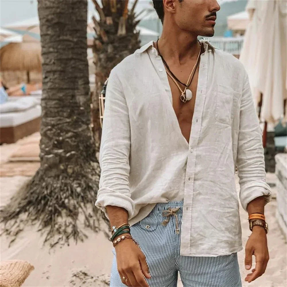 Uomo in camicia di lino bianca aperta e pantaloncini a righe blu su spiaggia tropicale, accessoriato con collane e bracciali. Moda estiva casual.