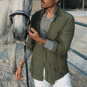 Uomo con camicia verde accanto a cavallo grigio in recinto. Moda casual maschile, equitazione, camicia di lino, pantaloni bianchi.