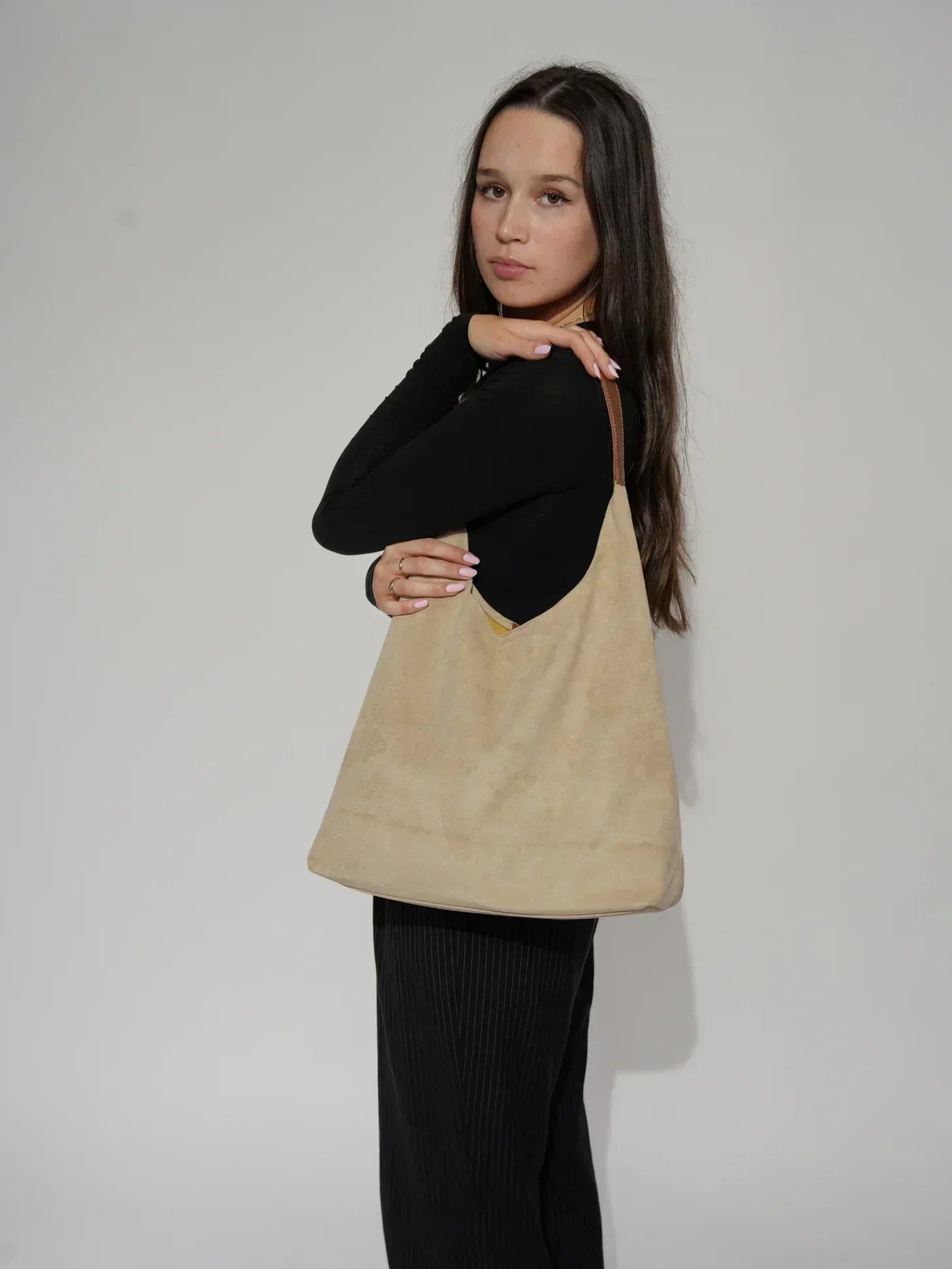 Donna con borsa a spalla beige in pelle scamosciata, manico marrone, indossa maglia nera e pantaloni scuri. Accessorio moda elegante e versatile.