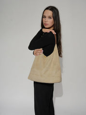 Donna con borsa a spalla beige in pelle scamosciata, manico marrone, indossa maglia nera e pantaloni scuri. Accessorio moda elegante e versatile.