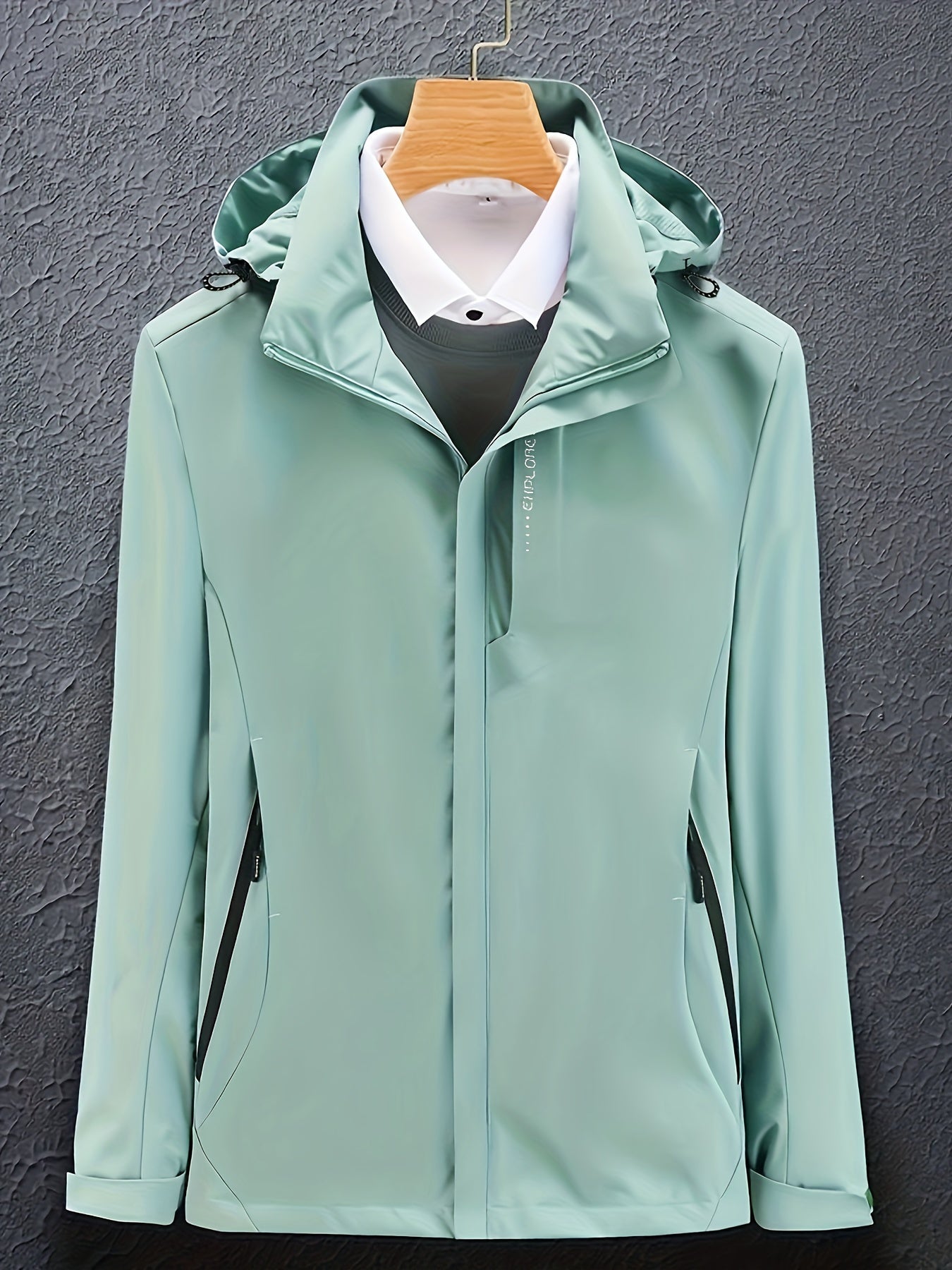 Giacca impermeabile verde menta con cappuccio, appesa su gruccia, su sfondo grigio. Design elegante, ideale per moda uomo autunno-inverno.