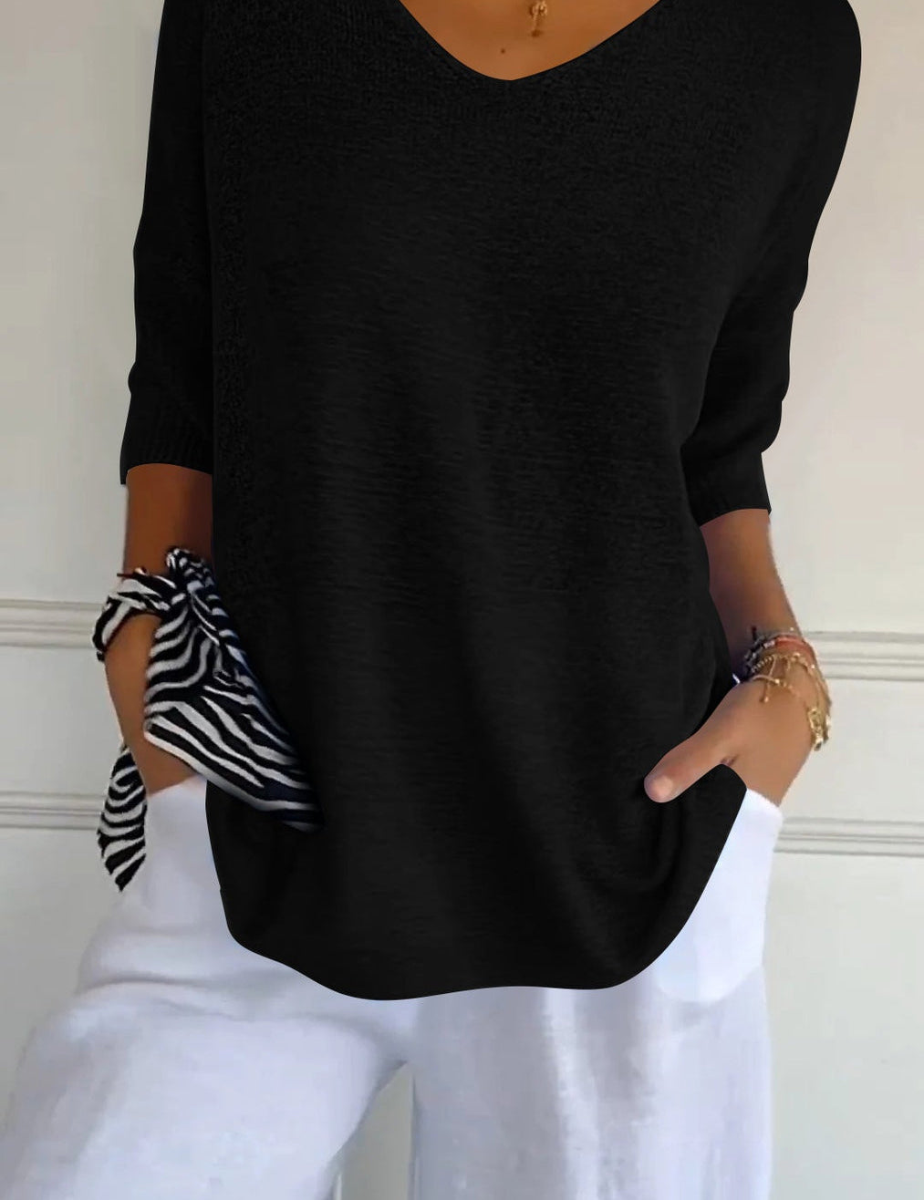 Donna con maglia nera a maniche lunghe, scollo a V, abbinata a pantaloni bianchi. Accessori: collana dorata, bracciali, foulard a righe.