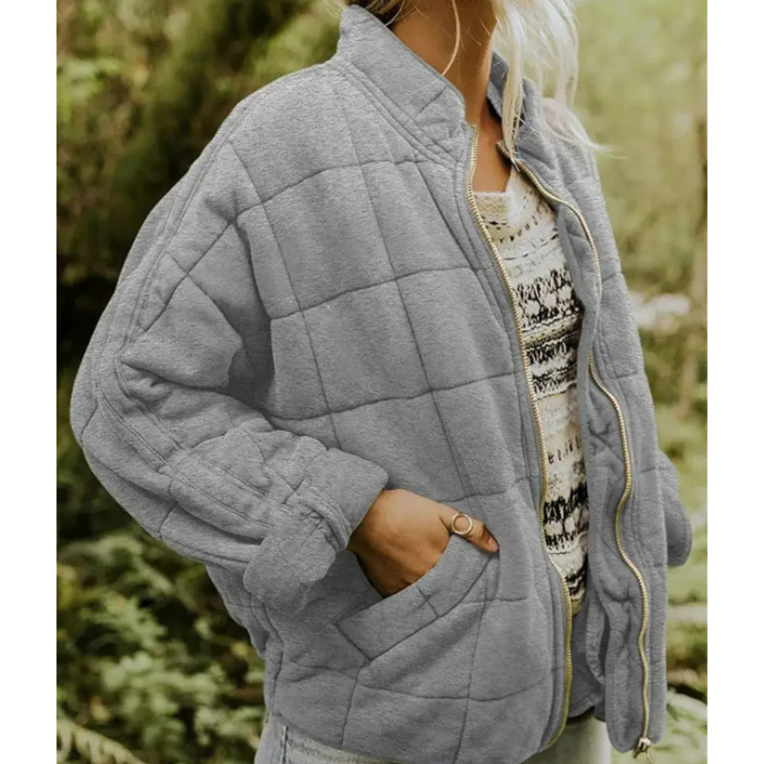 Giacca trapuntata grigia da donna, stile casual, con zip e tasche, indossata su maglia a fantasia. Perfetta per l'autunno e l'inverno.