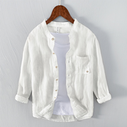 Camicia bianca in lino da uomo con colletto alla coreana, maniche lunghe e taschino, appesa su gruccia di legno. Elegante e casual.
