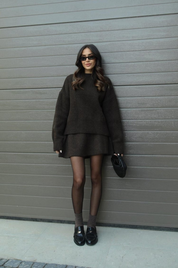 Donna con abito marrone, occhiali da sole e borsa nera, posa davanti a porta grigia. Look autunnale elegante, moda femminile contemporanea.