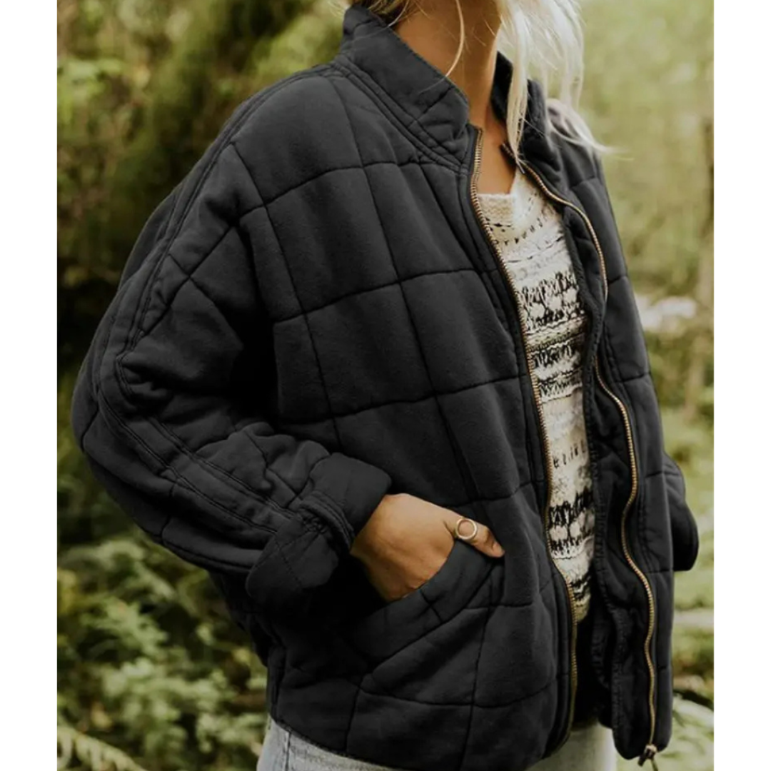 Giacca trapuntata nera da donna, stile casual, con zip frontale e tasche, indossata su maglia bianca a motivi, ambientata in un contesto naturale.