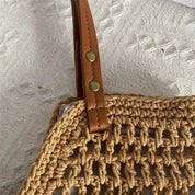 Borsa a rete in paglia intrecciata con manico in pelle marrone e bottoni metallici, su sfondo tessuto bianco. Accessorio moda estivo elegante.