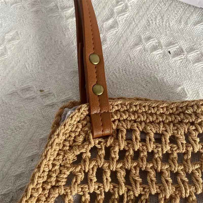 Borsa a rete in paglia intrecciata con manico in pelle marrone e bottoni metallici, su sfondo tessuto bianco. Accessorio moda estivo elegante.