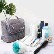 Borsa da toilette grigia su tavolo di marmo con prodotti per la cura personale: spazzola, lozioni, spazzolino, rasoio, bagnoschiuma, piante decorative.