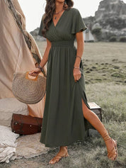 Abito lungo verde oliva con scollo a V e maniche corte, indossato da una donna all'aperto. Accessori: borsa di paglia, sandali. Moda estiva elegante.