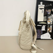 Zaino beige in tessuto morbido su tavolo, con tasche laterali e spallacci regolabili. Sfondo con bacheca nera e appunti. Accessorio moda casual.