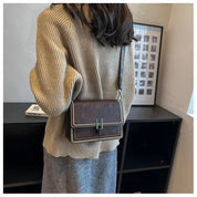 Donna con maglione beige e borsa a tracolla marrone in pelle, design elegante con cuciture a contrasto, su sfondo di interni moderni.