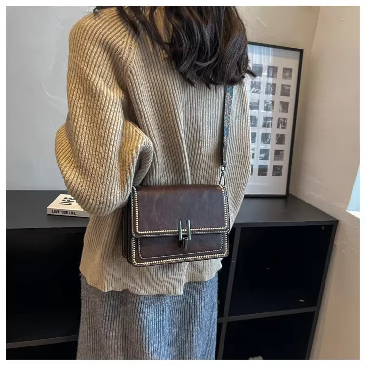 Donna con maglione beige e borsa a tracolla marrone in pelle, design elegante con cuciture a contrasto, su sfondo di interni moderni.
