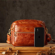 Borsa a tracolla in pelle marrone con tasche multiple, accanto a uno smartphone nero su tavolo di legno. Accessorio moda elegante e funzionale.