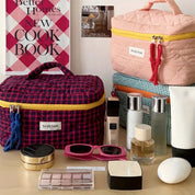 Set di borse da trucco colorate su tavolo con cosmetici, occhiali da sole e profumi. Sfondo con libro di cucina. Accessori moda e bellezza.
