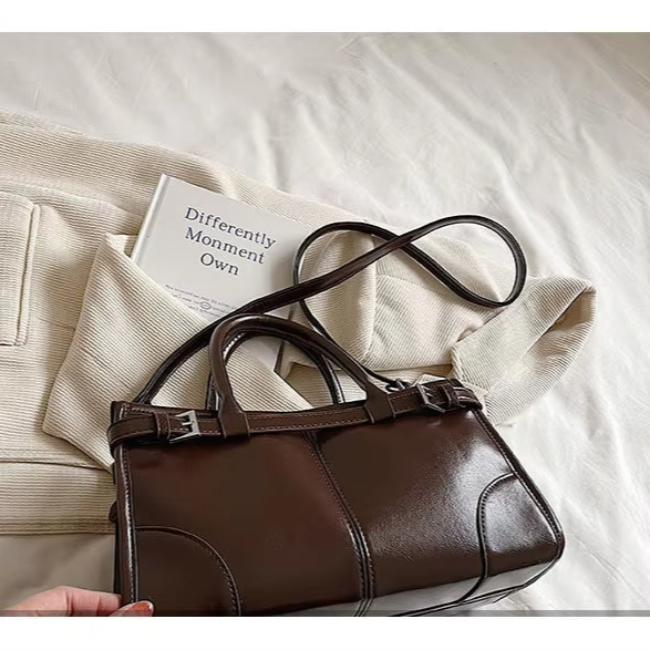 Borsa a mano in pelle marrone su letto bianco, accanto a libro e maglione beige. Elegante accessorio moda donna, stile classico e sofisticato.