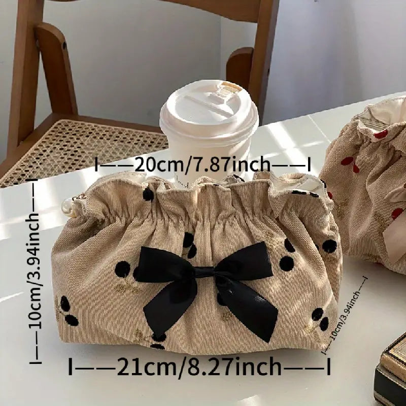 Borsa cosmetica beige a pois neri con fiocco nero, dimensioni 21x10 cm, accanto a bicchiere di caffè su tavolo in legno. Accessorio moda elegante.
