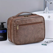 Borsa da toilette in pelle marrone con cerniera, manico superiore, accanto a lavandino e rasoio. Elegante organizer da viaggio per uomo.