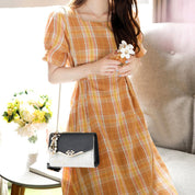 Donna con abito a quadri arancione, borsa a tracolla nera con dettagli dorati, seduta su divano. Moda estiva, accessori eleganti, stile casual.