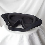 Maschera per dormire nera in memory foam su sfondo bianco, design ergonomico, cinturino regolabile, ideale per bloccare la luce e migliorare il sonno.