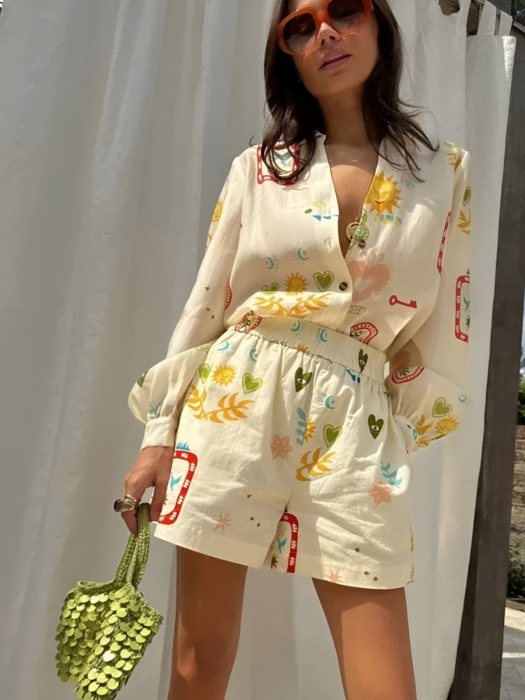 Donna con occhiali da sole indossa tuta estiva stampata con motivi floreali e geometrici, borsa verde a mano, sfondo bianco. Moda estiva femminile.