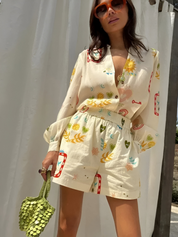 Donna con occhiali da sole indossa tuta estiva stampata con motivi floreali e geometrici, borsa verde a mano, sfondo bianco. Moda estiva femminile.